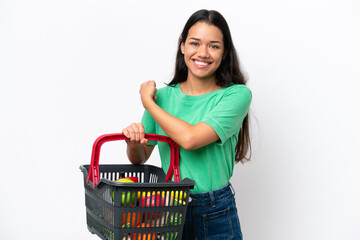 Young Colombian woman holding a shopping basket full of food isolated on white background pointing...