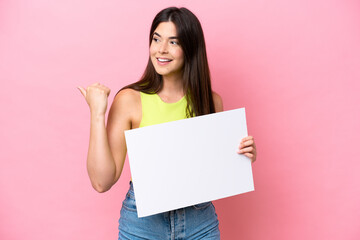 Young Brazilian woman isolated on pink background holding an empty placard and pointing side