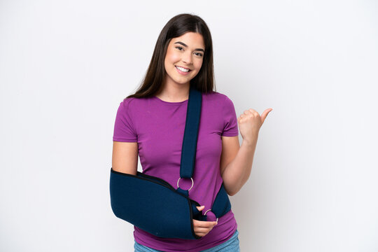 Young Brazilian woman with broken arm and wearing a sling isolated on white background pointing to the side to present a product