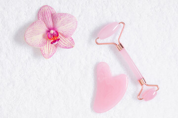 Pink jade roller and gua sha tool, orchid flower on white towel background. Flatlay of gua sha massage roller, stone scraper tool on a white background.