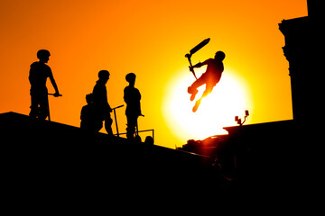 Unrecognizable teenage boy silhouette showing high jump tricks on scooter against orange sunset sky at skatepark. Sport, freestyle, extreme, youth, urban culture, outdoor activity concept