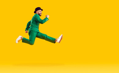 Fototapeta na wymiar Happy cheerful young man wearing a green suit and a black hat hurrying to a St Patrick's Day party, running fast and jumping high in the air isolated on a bright yellow color copy space background