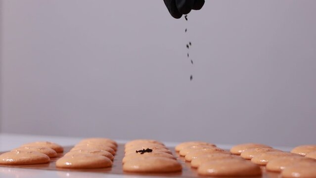 close up of process of making chocolate macarons. chef in black gloves is decorating brown shells of Macaroons and throwing black sesame on it. slow motion video.