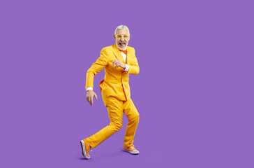 Active life and leisure of older people. Cheerful trendy, eccentric and energetic senior man funny dancing isolated on purple background. Full length of funny male retiree in yellow suit and sneakers.