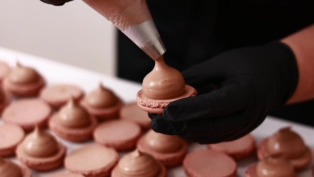 Making french desert macarons. Chef hands in black gloves with confectionery bag add cream to the ready halves of brown chocolate macaroons at pastry shop. cooking, food and baking concept