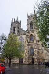 View of York Minster cathedral building exterior during rain in morning with people walking on pavement and cloudy sky background.