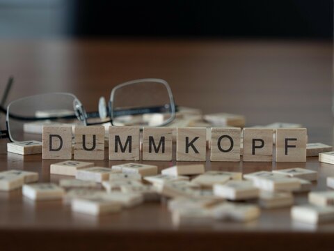 dummkopf Wort oder Konzept dargestellt durch h&ouml;lzerne Buchstabenfliesen auf einem Holztisch mit Brille und einem Buch