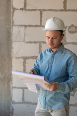engineer in a white helmet with a project in his hands is thoughtful and professional. A successful European constructor or builder at the facility makes measurements of the room and takes notes. Male