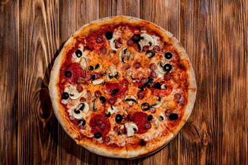 Tomato-based pizza with mozzarella cheese, hunting sausages, salami, olives, mushroom sauce. On a wooden background