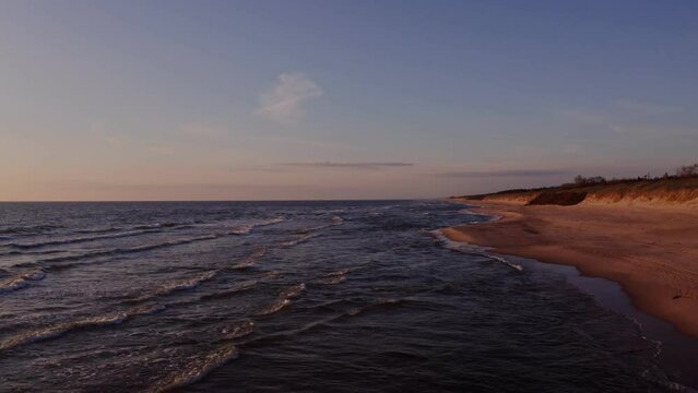 Panoramic view of a deserted beach washed by sea waves, turning into a view of the sea distance with the rising sun reflected in the sea water. Video from a drone.