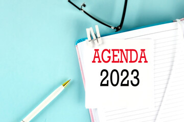 AGENDA 2023 text on a sticky on notebook with pen and glasses , blue background