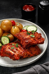 Dish, plate with Marinated pork bbq ribs, young potatoes, tomatoes on black dark background. Hearty Dinner, lunch, meal.