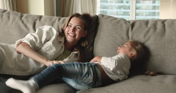Happy playful mom tickling excited screaming little daughter kid on couch, hugging, cuddling girl, laughing, giggling. Sitter playing active games with child at home. Family fun concept