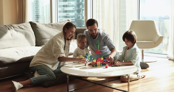 Happy young parents helping two sweet little kids to stack toy tower from building blocks, constructing colorful city model on table in living room, talking, laughing, playing learning game
