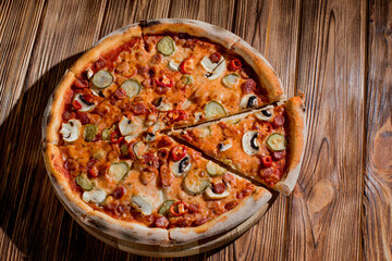 Appetizing pizza with hunting sausages, mozzarella cheese, pickled cucumbers, chili peppers on a tomato base on wood background