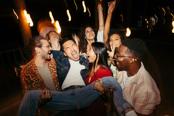 Multiracial friends carry a man at the open-air rooftop night party. - 526390937