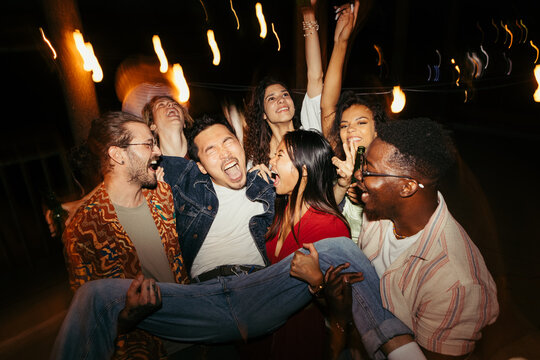 Multiracial friends carry a man at the open-air rooftop night party.