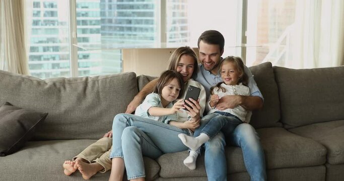 Cheerful little sibling kids and happy young parents taking selfie photo on cellphone on home couch. Family couple hugging, holding children in arms, using mobile phone for self picture