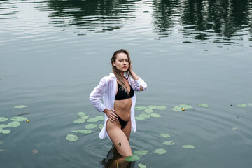 Gorgeous sensual blonde girl with wet hair in a white shirt and black swimsuit posing in the lake after the rain.