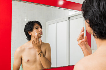 young man putting cream on his face while looking at himself through a mirror