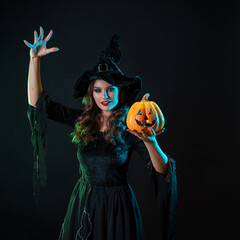 A young beautiful witch in a conical hat holds a Halloween pumpkin in her hands and makes a spell with her hand. Halloween Costume Party, Witches' Sabbath