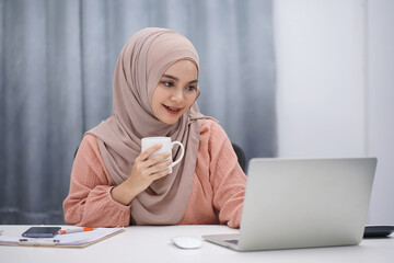 Beautiful asian muslim woman brown hijab drinking coffee and making a phone call while working at...
