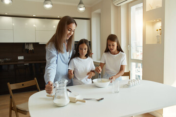 Obraz premium Happy family preparing food together in the kitchen. Mom teaches her daughters how to cook and knead the dough. Mother's Day concept. break eggs