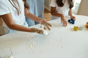 two little girls and their beautiful mother in aprons play and laugh while kneading dough in the...