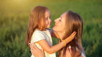 Young daughter shows pure love hugging mom on green meadow at sunset. Mother touches preschooler girl long hair and hugs daughter closeup, sunlight