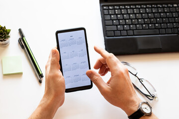 Human hand touching on screen of smartphone. Mobile phone with an open calendar for 2022 year in a male hand on a working desk. On the desktop a laptop, two pens, glasses, dairy