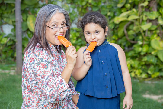 Happy Indian Senior Woman Having Ice Lolly Or Ice Cream With Little Girl Child Together Outdoor, Old Mature People Enjoy Retirement Life. Summer Holidays, Grandmother And Granddaughter Spend Time.