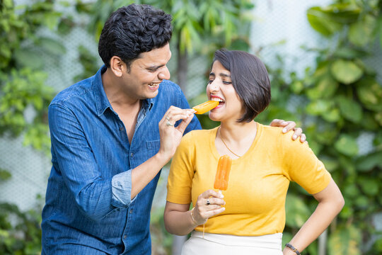 Happy Young Indian Couple Enjoying Ice Lolly Or Ice Cream Together Outdoor, Love And Bonding Of Husband Wife.