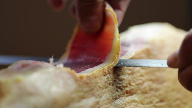 熟成された生ハム原木を、専用のナイフで薄くスライスカットする動作。透き通るような身を丁寧に切り出す職人技を捉えたクローズアップ映像。飲食店、バル、高級食材やワインをテーマにした広告映像に。 | Slicing a block of cured ham with a knife.