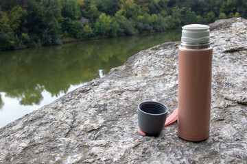 Beige thermos bottle and cup of tea or coffee on rock over the river. Beautiful summer, spring, autumn landscape. Picnic on the rock - obrazy, fototapety, plakaty