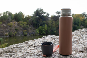 Beige thermos bottle and cup of tea or coffee on rock over the river. Beautiful summer, spring, autumn landscape. Picnic on the rock