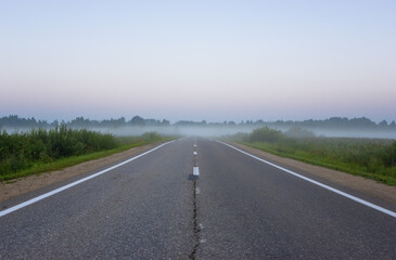 Empty, straight suburban asphalt highway with white road markings vanishing into the morning mist at dawn. Yaroslavl Oblast, Russia. - 526436779