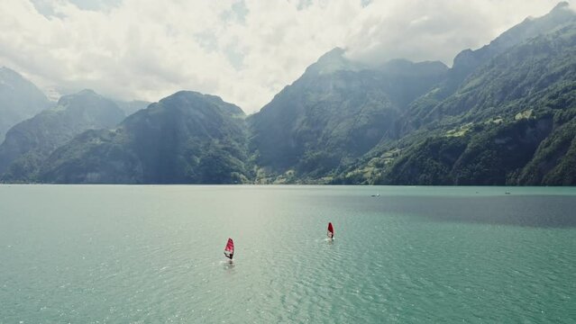 Two men are windsurfing on a picturesque lake at the foot of the Alps in Switzerland. Drone point of view. The tops of the mountains are covered with thick white clouds