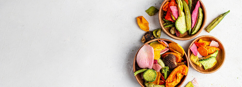 Dehydrated vegan chips crisps in wooden mango bowl. Vitamin salty free healthy fast food with carrot slices, beetroot wedges, broccoli, zucchini on a light table. food photo banner copy space top view
