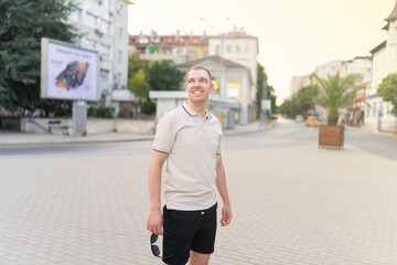 Fototapeta premium Varna,Bulgaria - 15.08.22 - Young stylish man wearing t-shirt with beard in glasses, standing on the street on city background.