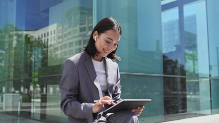 Asian elegant professional business woman wearing suit using digital tablet outside office, happy businesswoman executive holding pad corporate technology device on city street glass background. - Powered by Adobe