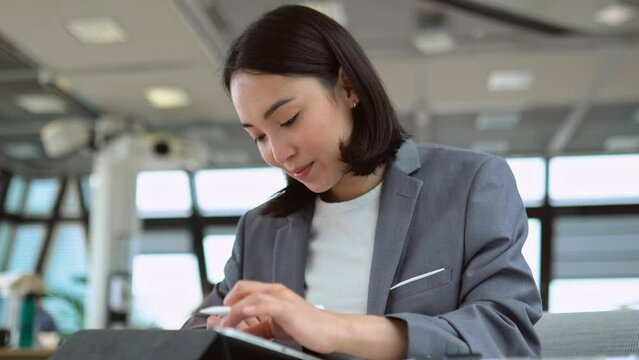 Young professional Asian business woman employee marketing manager wearing suit using digital tablet signing e document, working on online corporate management in big modern coworking office space.