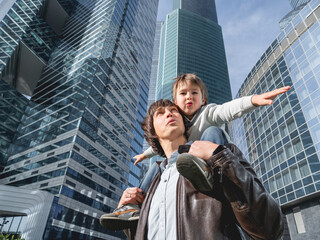 Laughing boy plays like airplane on father's shoulders. Dad and son looks on glass walls of buildings. Future and modern technologies, life balance and family life in well keeps districts.