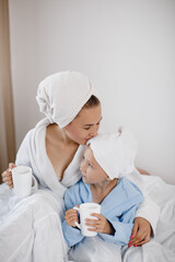 Mom hugs and kisses her little daughter in bed in the morning. Mom and her little daughter in bathrobes and towels on their heads drink tea in bed. Family concept. 