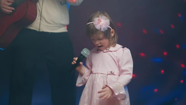 Little girl on stage in vintage dress, she sings into microphone and dances, her father plays acoustic guitar. Color music is shining and smoke is billowing. Father and Child Day, performance on stage