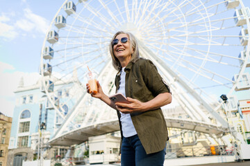 Happy grey haired European woman enjoying traveling, holding phone, smartphone while having juice on the go using plastic cup wearing sunglasses and green shirt in city and ferris wheel on background