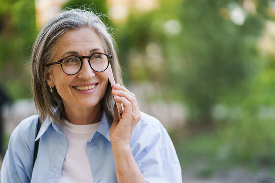 Senior grey hair woman in eye glasses smiling talking on the phone standing outdoors garden or park background wearing blue shirt. Silver hair woman outdoors