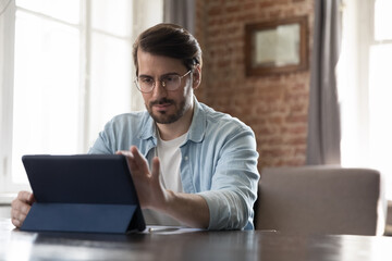 Focused millennial freelance employee man in casual using tablet computer, working with online app, sitting at workplace table in loft home office, touching screen. Gadget user using Internet service