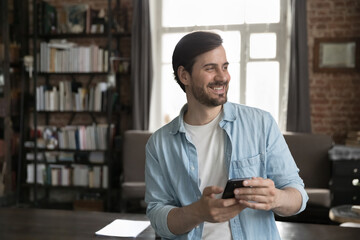 Obraz na płótnie Canvas Cheerful dreamy millennial smartphone user man holding cell, standing at work table in home office, looking away with happy smile, feeling inspired, thinking, enjoying wireless online communication