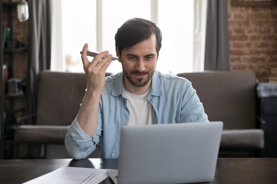 Positive freelance employee man listening audio from smartphone at home office workplace. Businessman in casual using professional online app on laptop, receiving voice message on mobile phone - Powered by Adobe