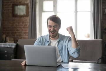 Excited millennial freelance business man getting good news message, email, notice, celebrating project success, sitting at laptop with happy smile, making winner hand, laughing, shouting